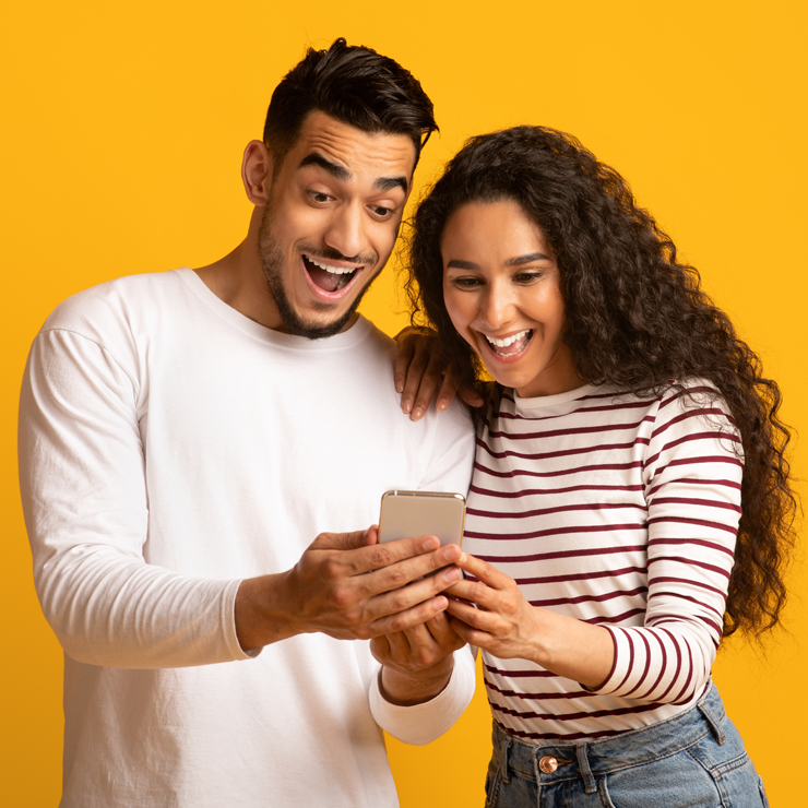 Homem e mulher brancos posam segurando celular sob fundo laranja.